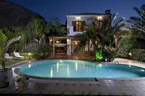 Una piscina con zona de patio, palmeras y una casa con techo de tejas.