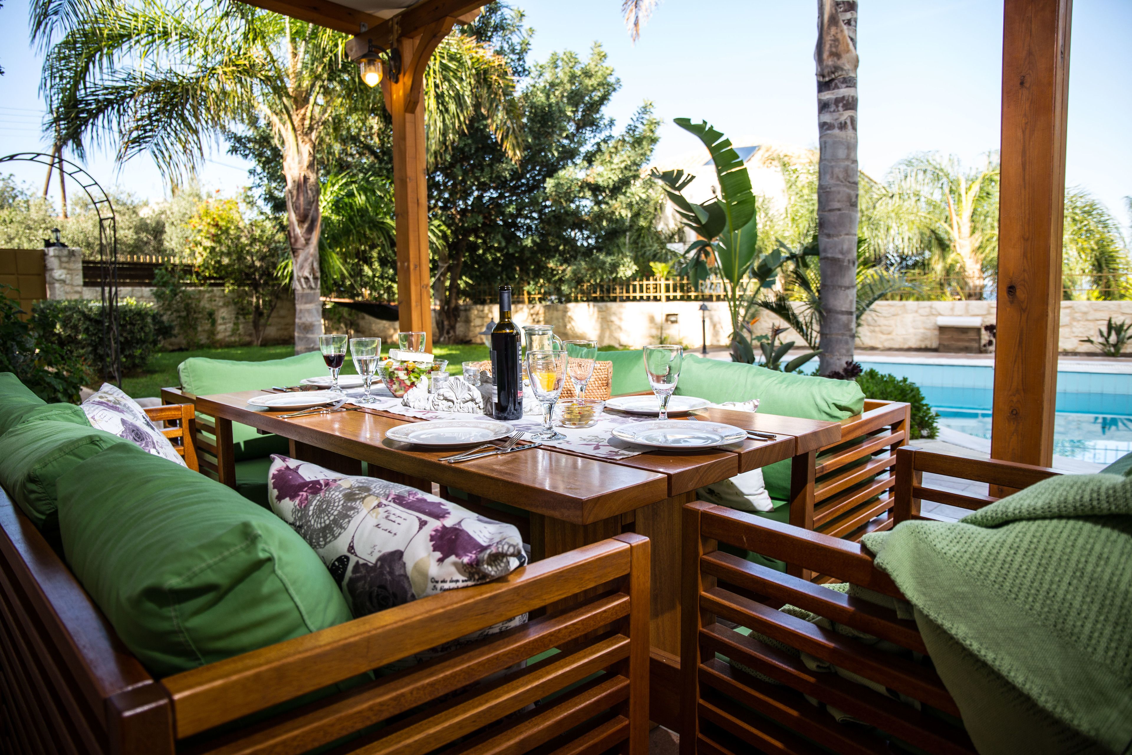 Un espace de restauration en plein air, doté de mobilier en bois et de coussins verts, offrant une vue sur une piscine et entouré d’une végétation luxuriante.