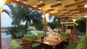 An outdoor patio with wooden furniture, a table set with a bottle and glasses, and a swing chair. - Pandora Villas (Hersonissos)