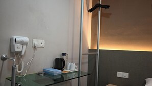 Habitación de hotel con repisa de vidrio que sostiene un secador de pelo, una tetera y una caja. Se observa una cama con un almohadón y un control remoto sobre una mesita de noche de madera.