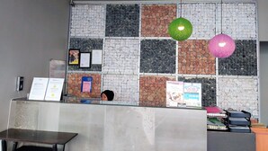 Un mostrador de recepción con una pared de azulejos al fondo, una persona sentada detrás y dos lámparas colgantes.
