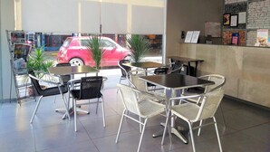 Un café con mesas y sillas, un amplio ventanal y un coche rojo en el exterior.