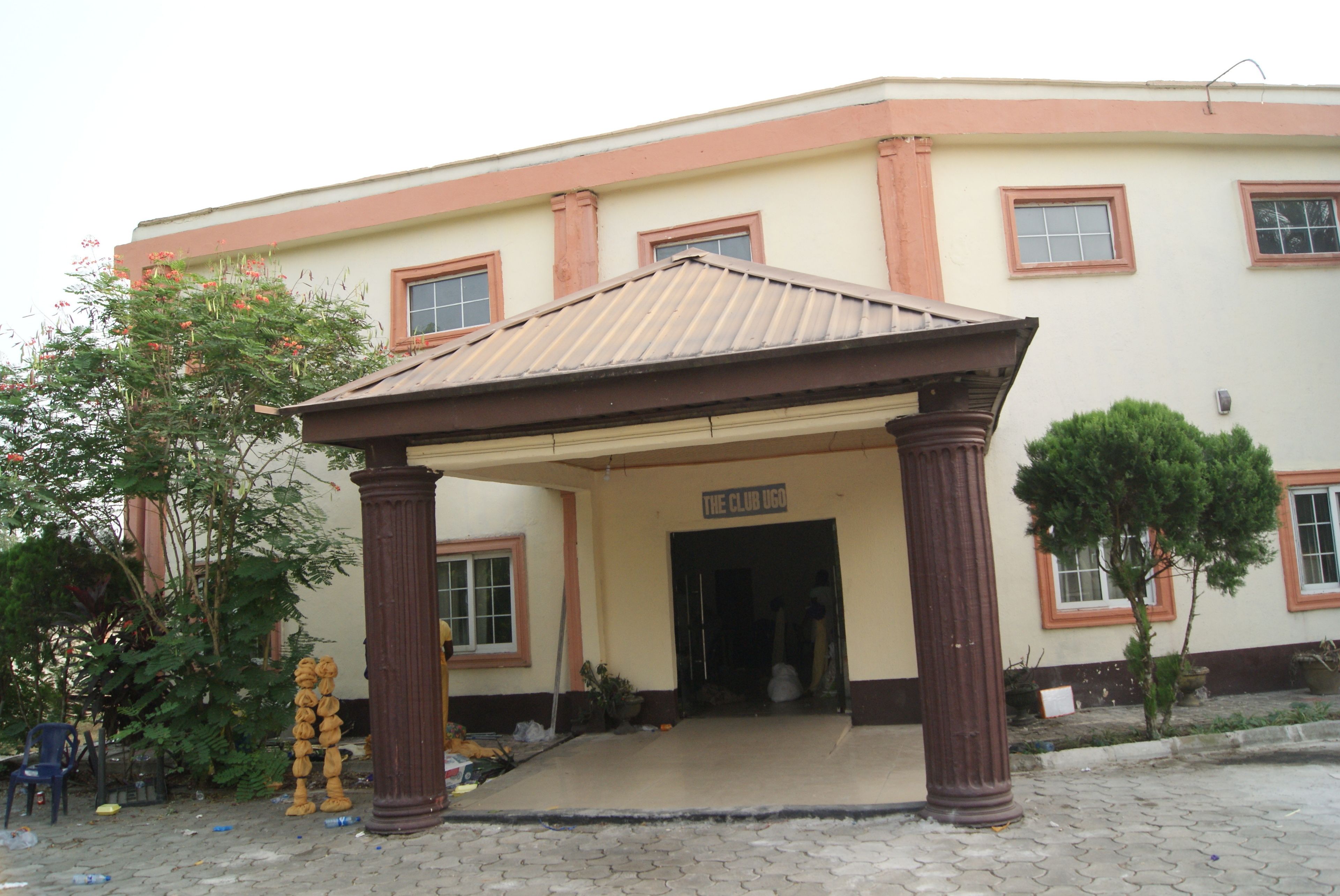 A building with a covered entrance, a sign reading "THE CLUB UGA", and a paved area with trees and chairs.