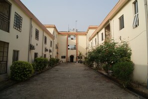 A courtyard with a paved walkway flanked by buildings with beige walls and red trim. There are small bushes along the sides and a tree with red flowers in the foreground.
