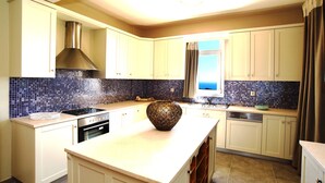 A kitchen with white cabinets, a stainless steel range hood, a blue-tiled backsplash, and a large island with a decorative bowl. - Liogerma Vacation Home (Kefalonia)