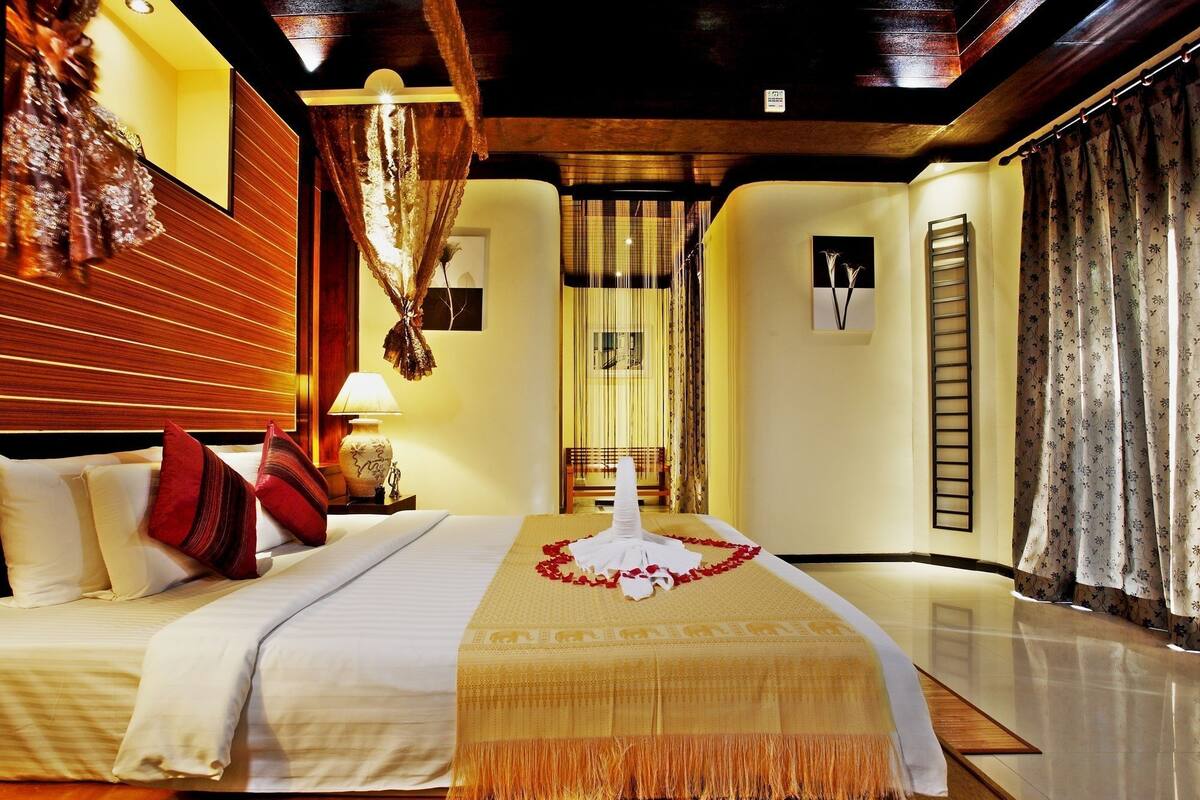 Una habitación de hotel con una cama bien hecha, un cabecero de madera, una mesita de noche con lámpara y un ventanal con cortinas.