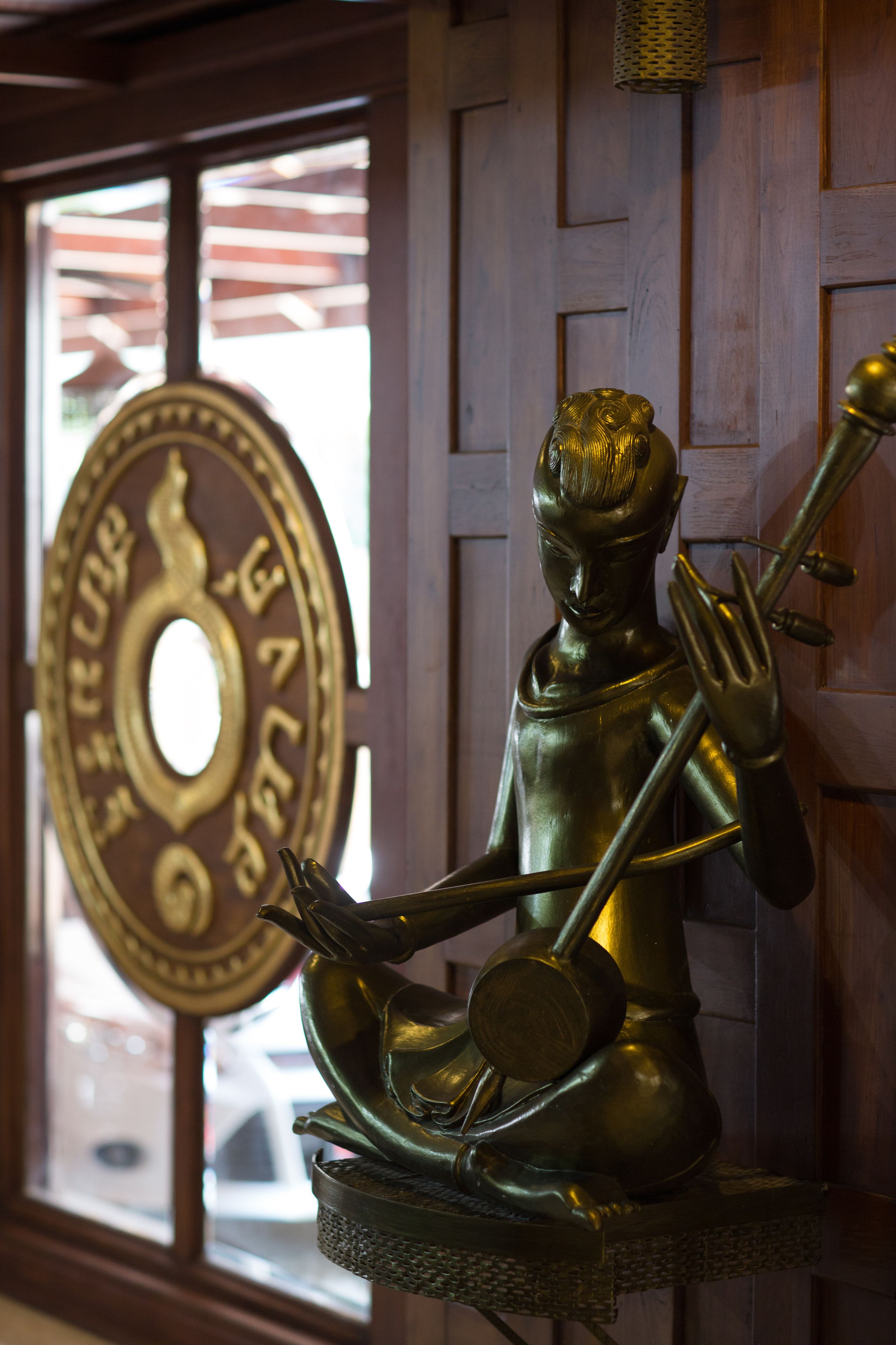 A bronze statue of a seated figure holding a staff and a bowl, positioned in front of a wooden paneled wall with a circular emblem.