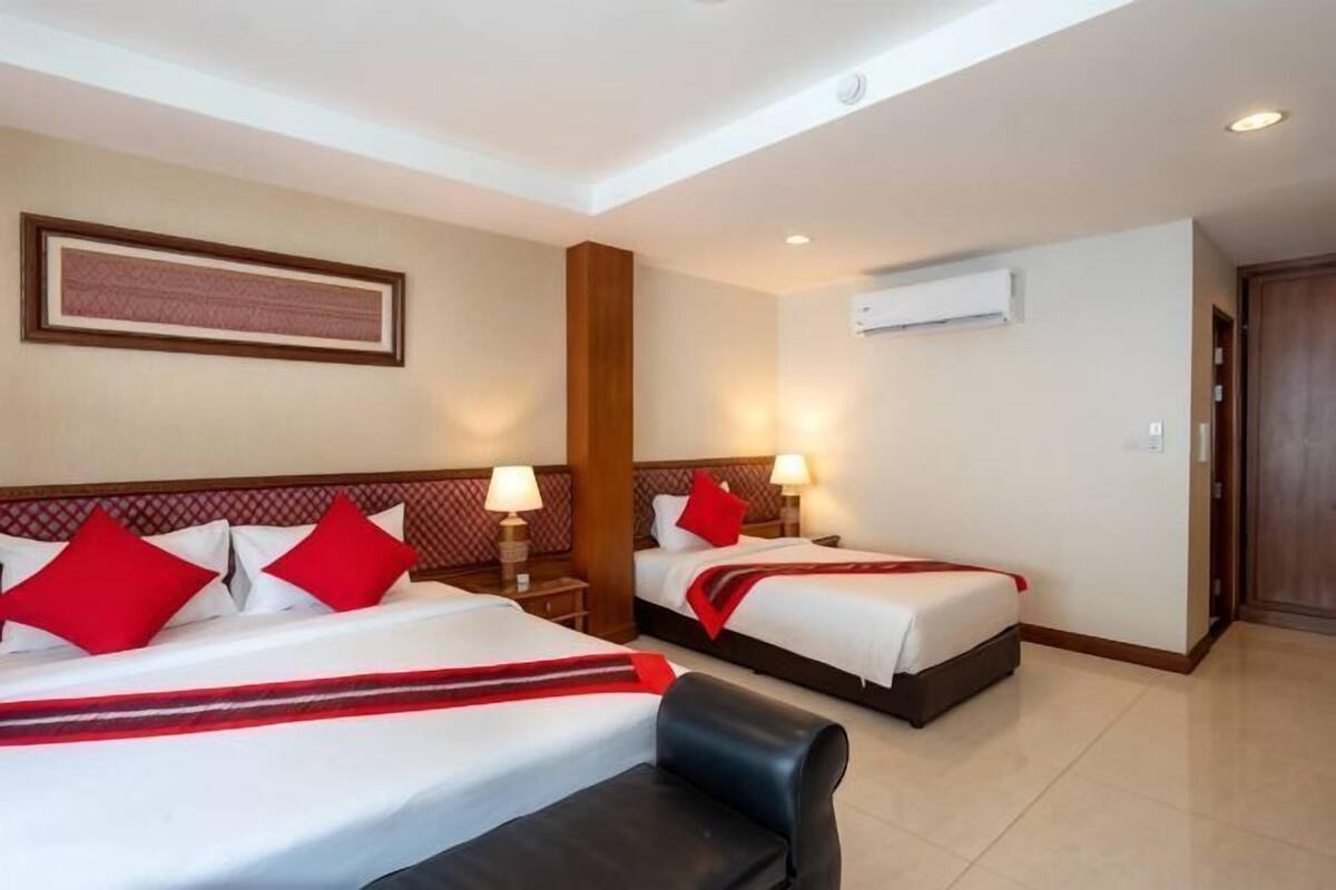 A hotel room with two beds, a black sofa, a wooden headboard, a framed picture on the wall, and an air conditioning unit.