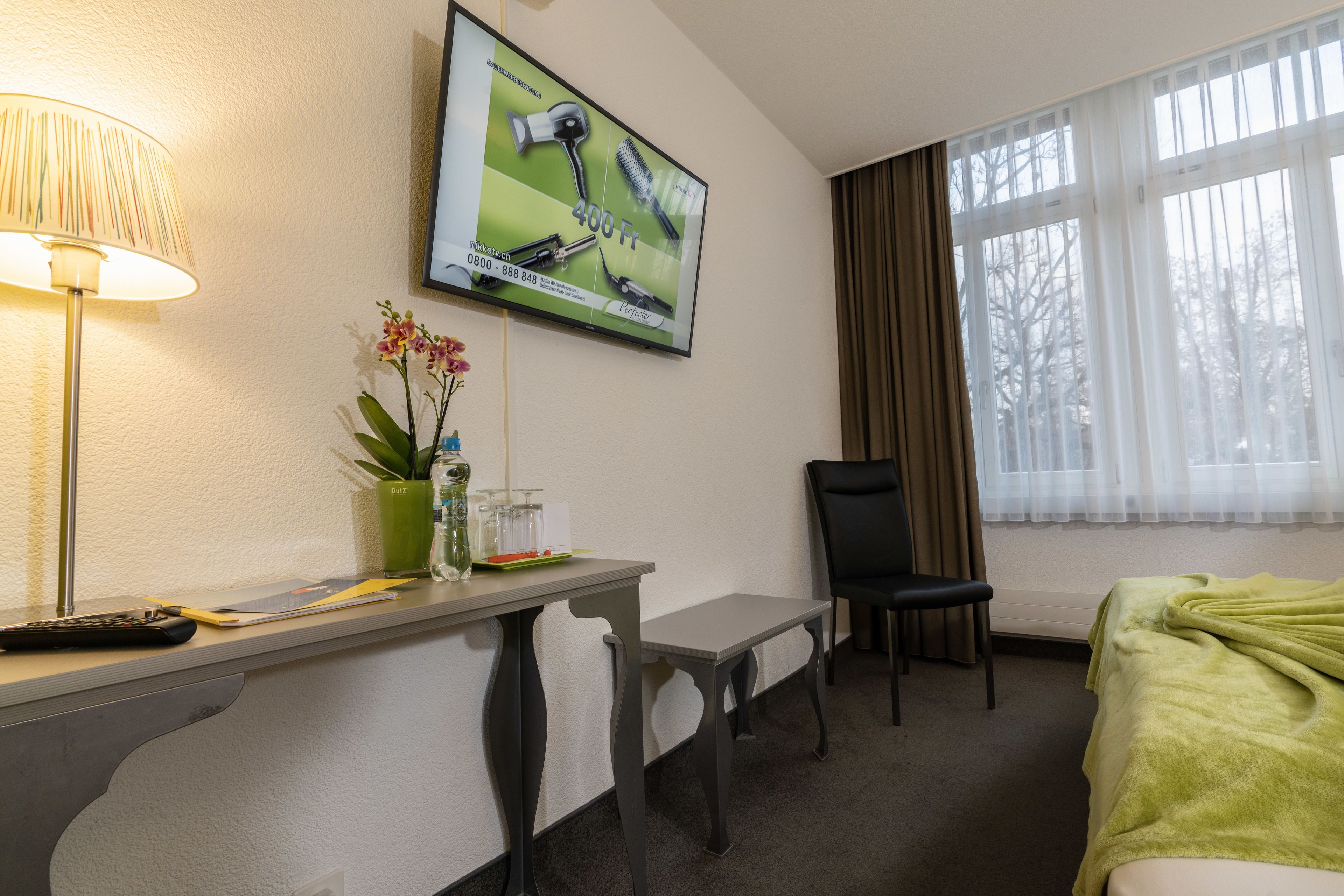 Una camera d'albergo con un letto, una scrivania con una lampada, una sedia, una televisione a parete e una finestra con tende.