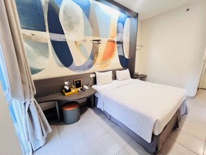 Une chambre d’hôtel moderne avec un grand tableau abstrait au mur, un lit soigneusement fait, une table de chevet avec une lampe et un petit tabouret rond.
