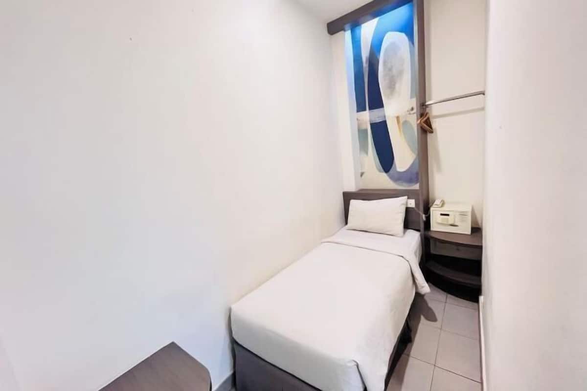 Una habitación pequeña con una cama individual, colcha blanca, una mesita de noche con un teléfono y un cuadro colgado en la pared.