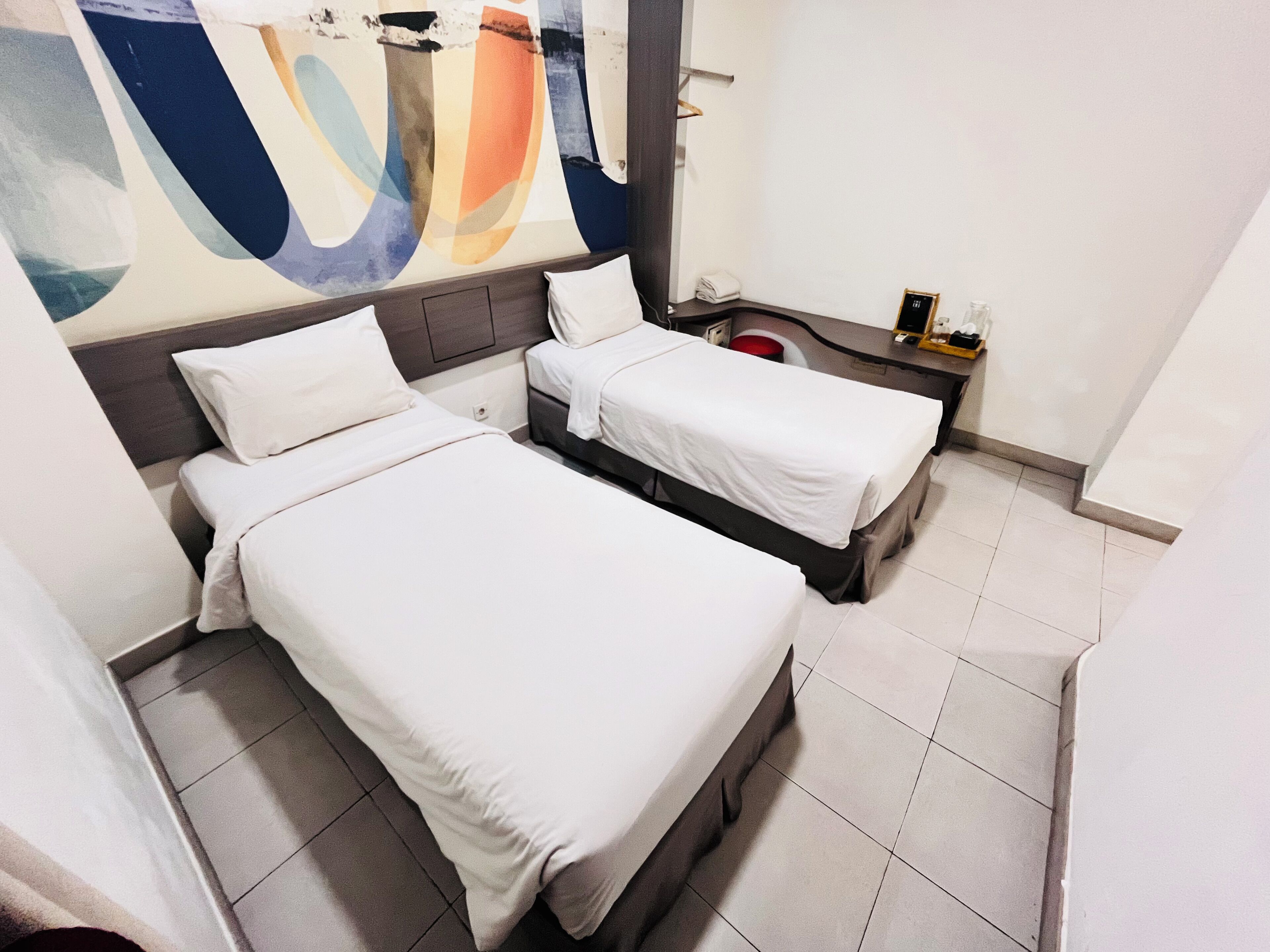 Habitación de hotel con dos camas individuales, un cuadro abstracto grande en la pared y una pequeña mesa con un recipiente rojo.