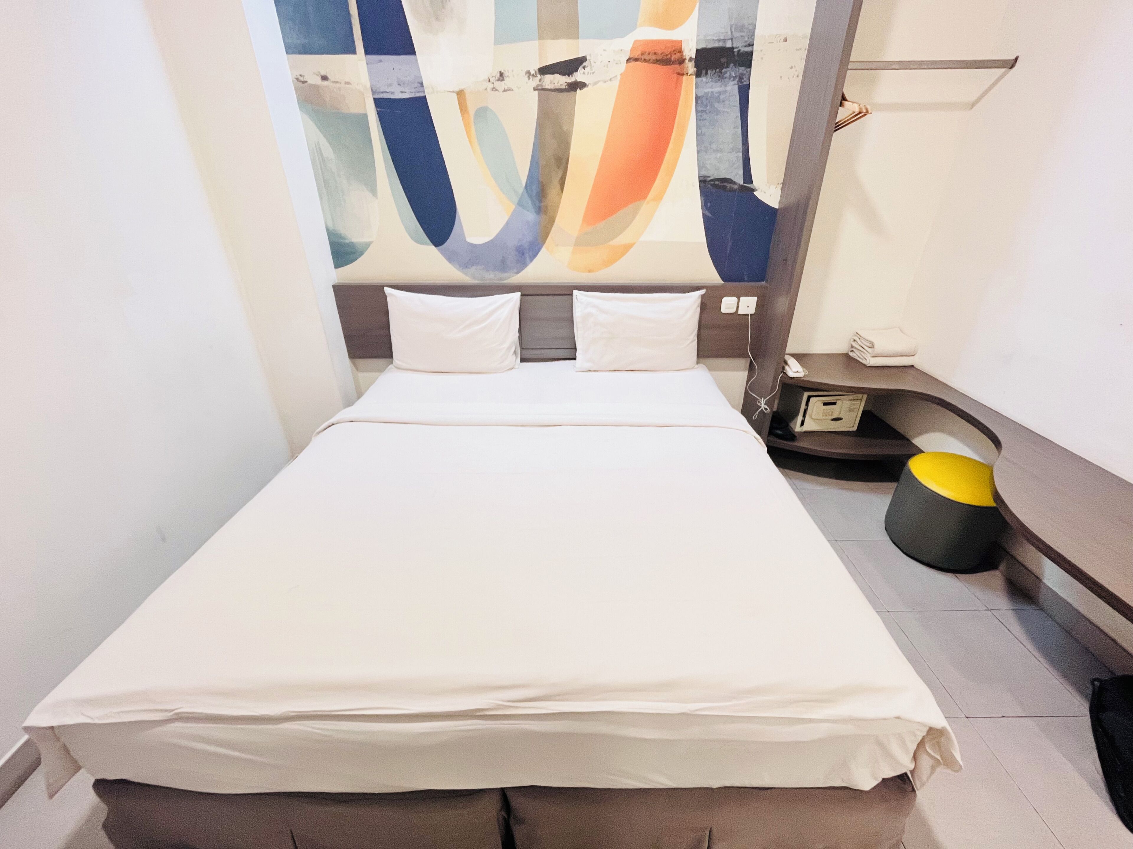 Ein modernes Hotelzimmer mit einem großen Bett, einem Nachttisch, einem kleinen Hocker und einem Wandbild.
