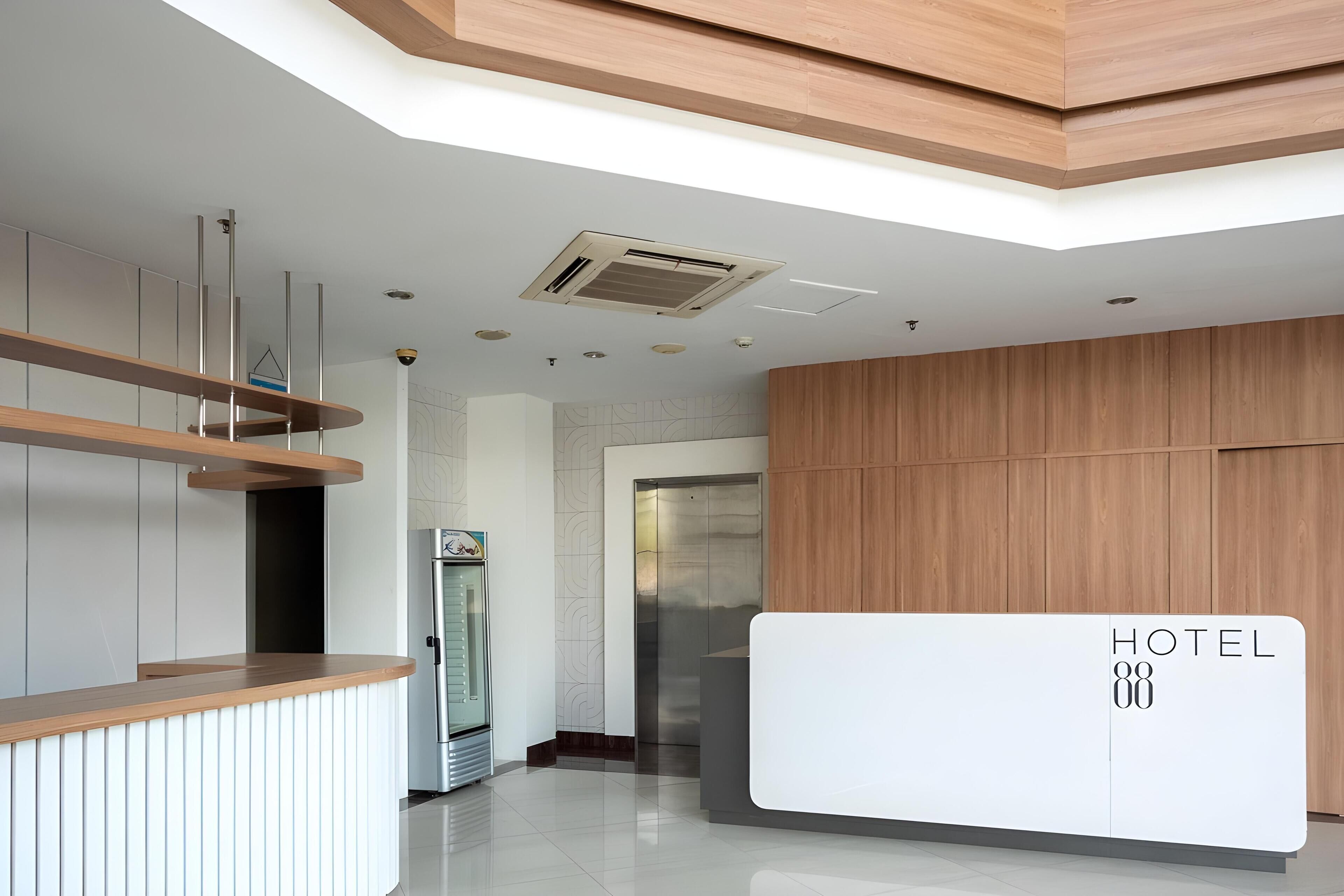 A modern hotel reception area with a wooden reception desk, a glass elevator, and a stainless steel refrigerator.