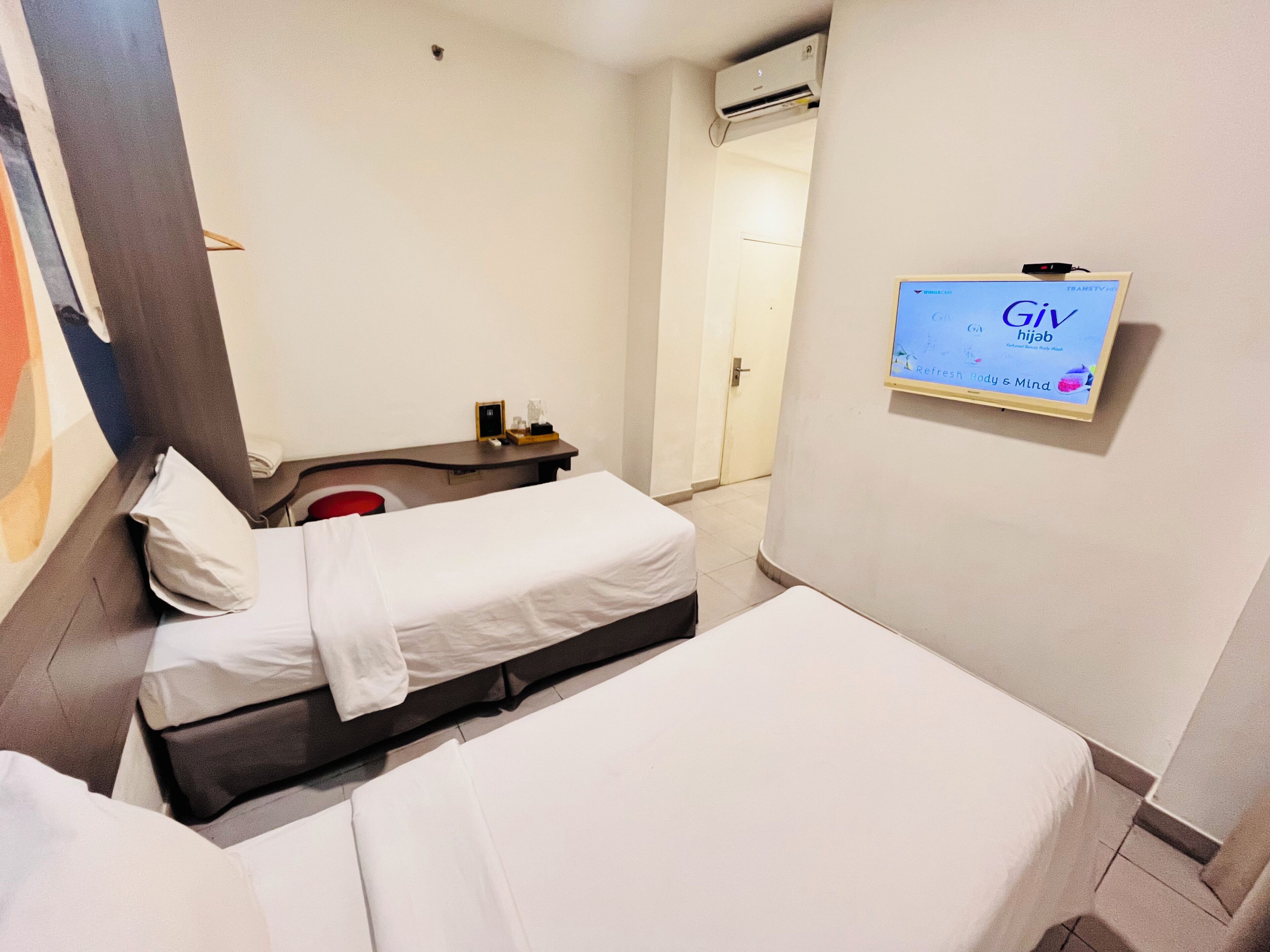 A hotel room with two single beds, a small desk with a chair, a wall-mounted TV displaying 'Giv hijab', and an air conditioning unit on the wall.
