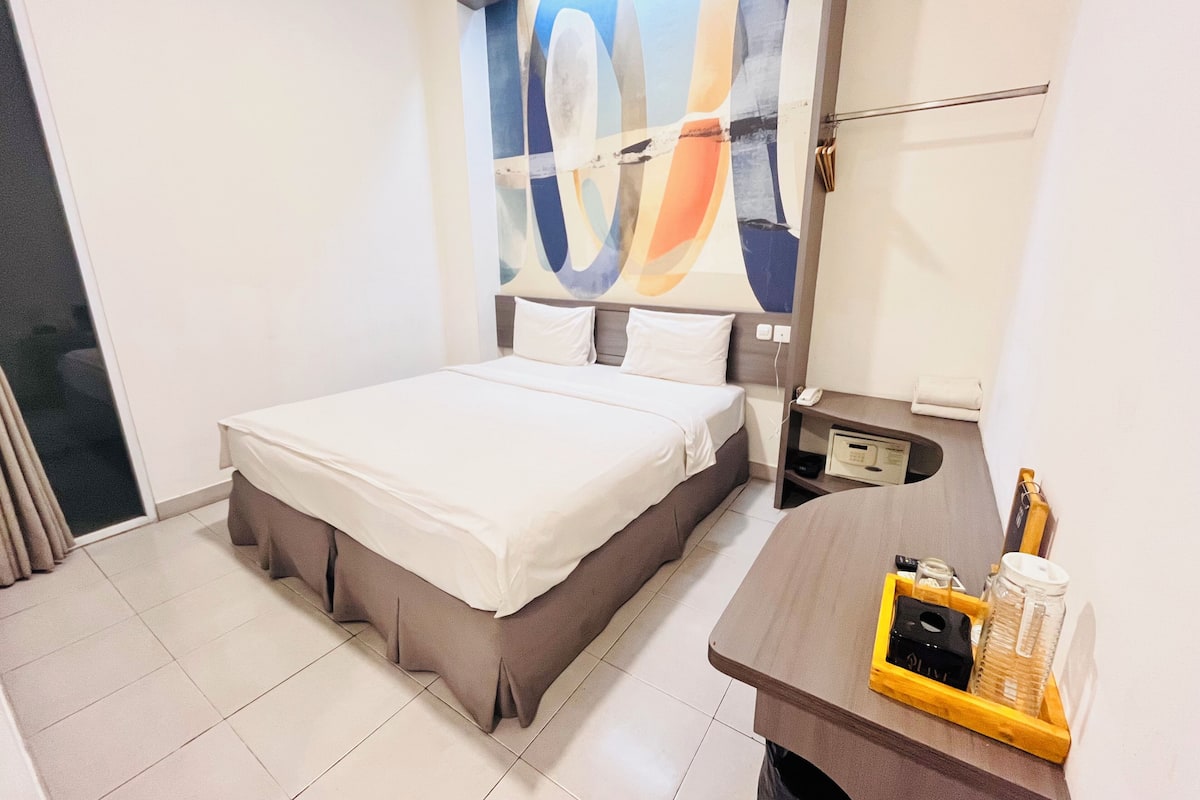 Una habitación de hotel moderna con una cama grande, una mesita de noche con un teléfono y un estante pequeño, además de un cuadro abstracto y colorido en la pared.