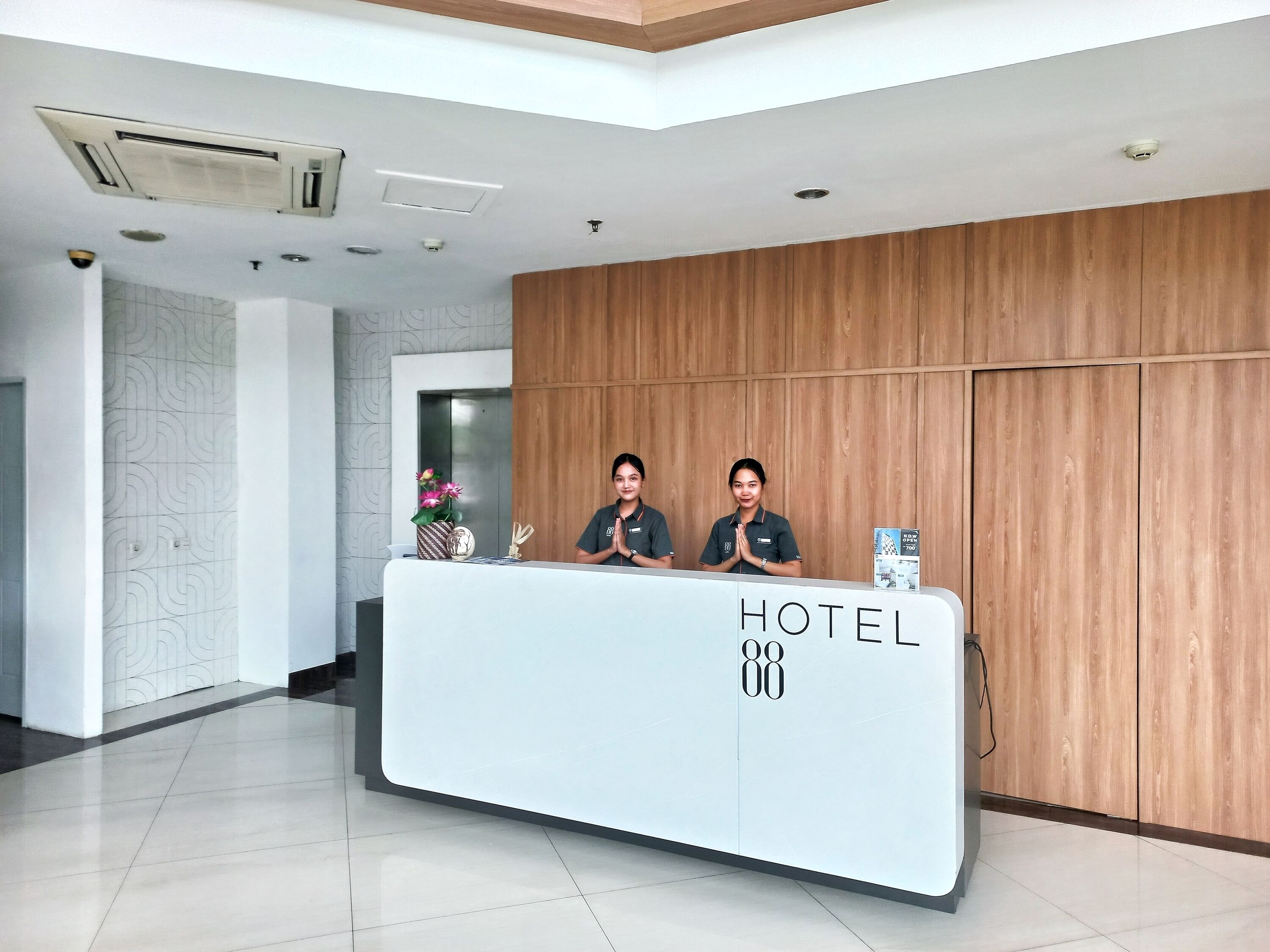 A hotel reception desk with two staff members, a modern interior, and a wooden panel wall.