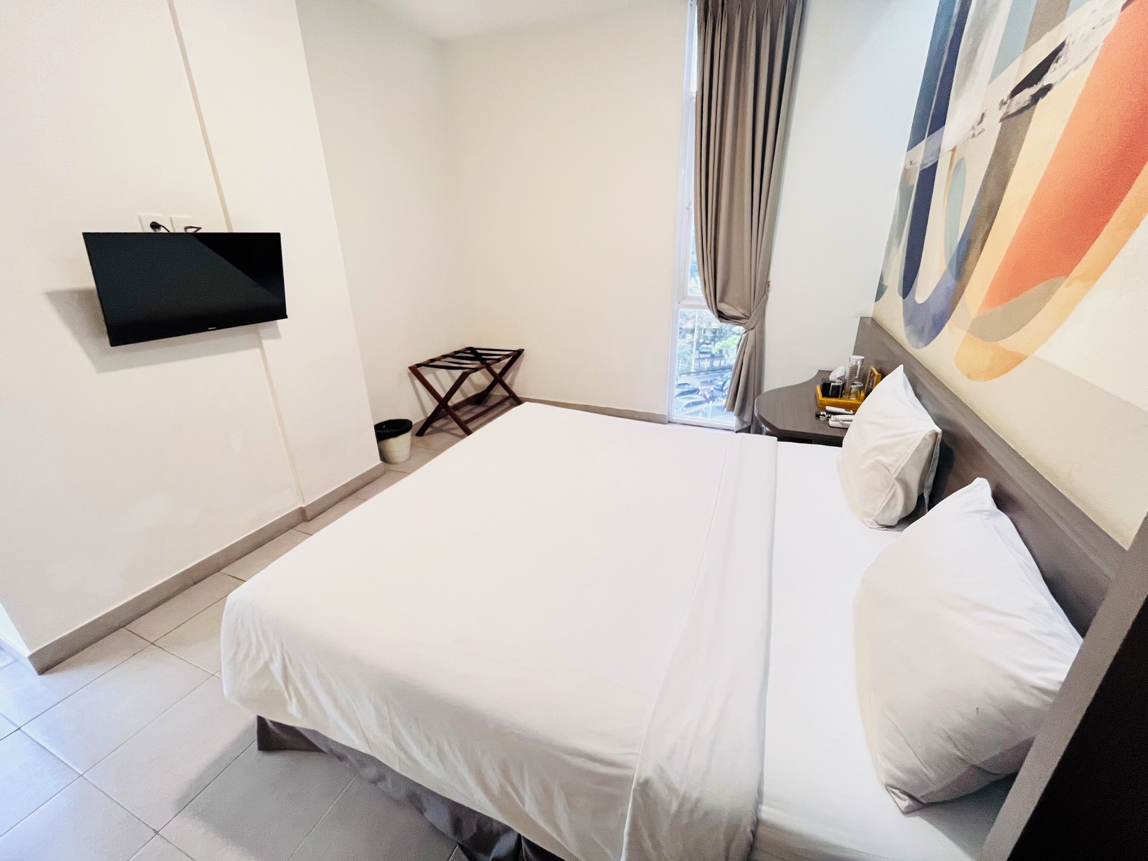 A hotel room with a bed, a TV mounted on the wall, a window with curtains, and a small table with a bottle and glasses.