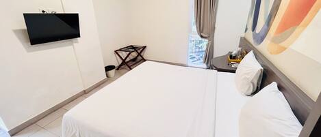 Habitación de hotel con cama, televisión montada en la pared, ventana con cortinas y una mesita con una botella y vasos.