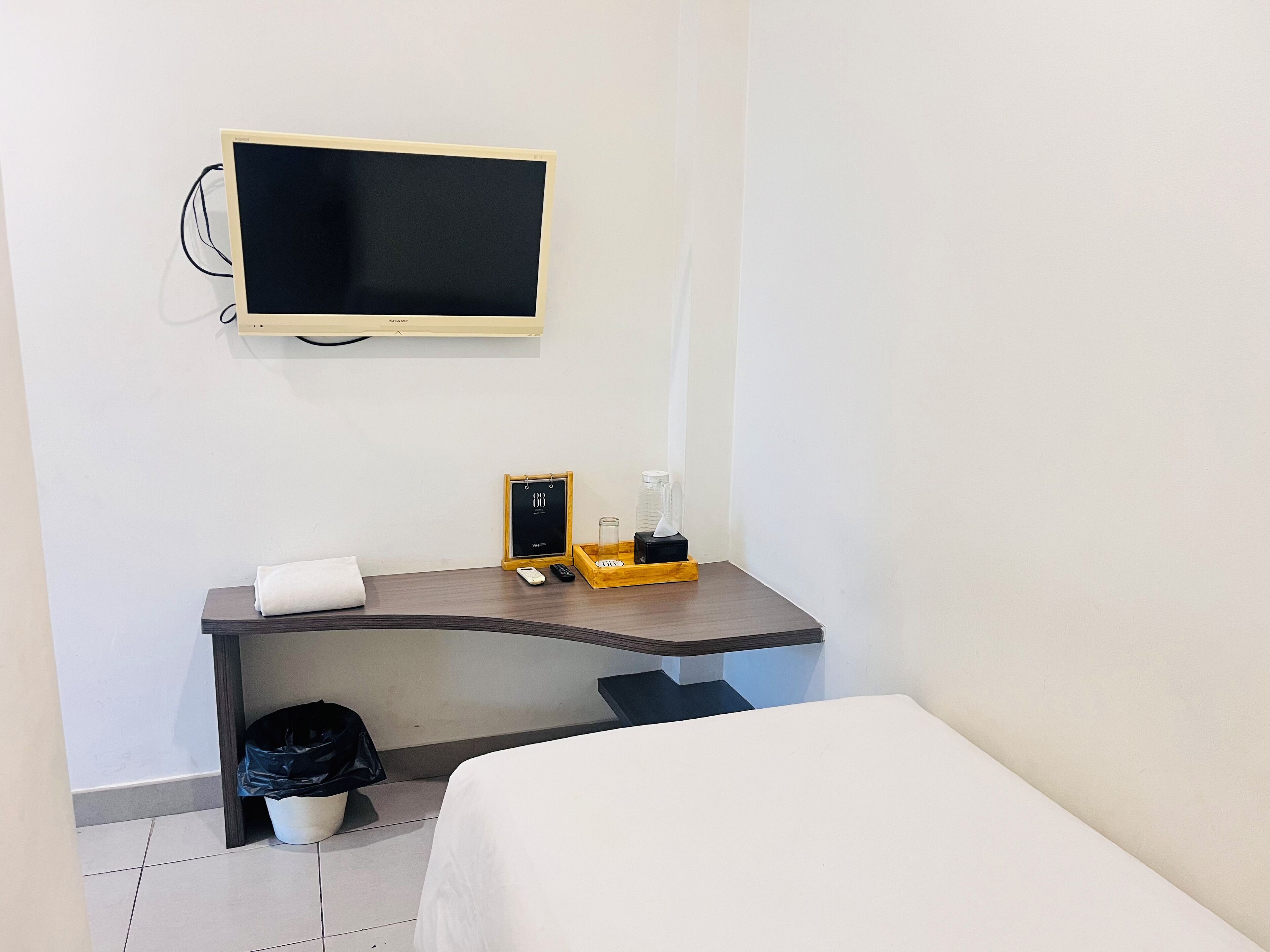 Ein Hotelzimmer mit Bett, Schreibtisch mit Stuhl, Wandfernseher, Mülleimer und einem kleinen Tisch mit Glas und Bilderrahmen.