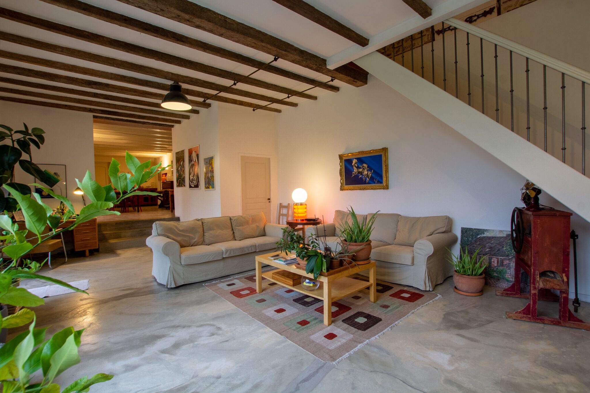 A living room with a sofa, coffee table, and potted plants. There is a staircase with a railing and a door leading to another room.