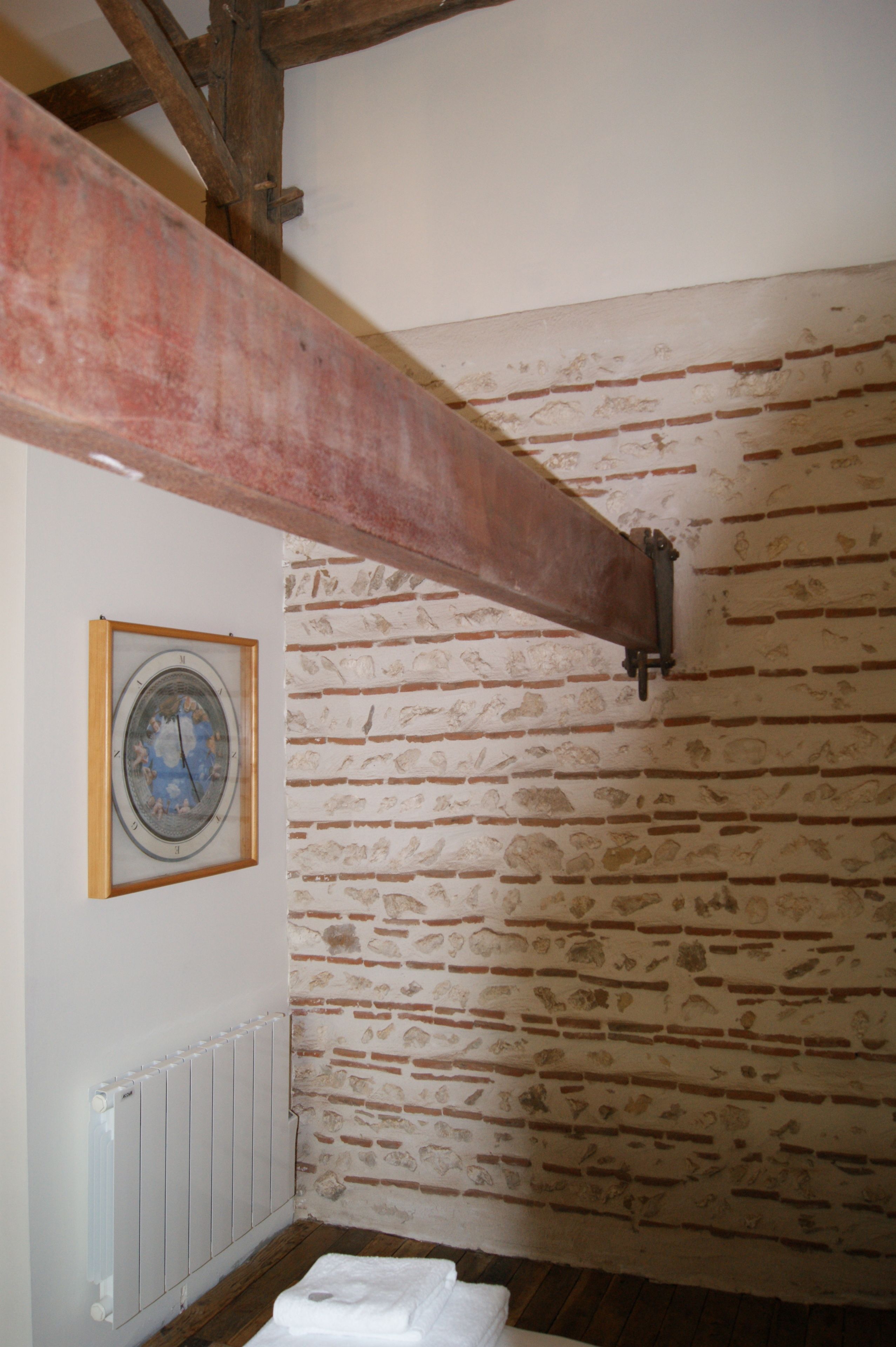 Une pièce aux murs en briques apparentes, avec une poutre en bois au plafond, un radiateur et une horloge encadrée accrochée au mur.