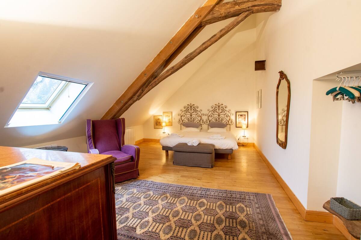 A cozy attic bedroom with a sloped ceiling, a wooden desk, a purple armchair, a bed with a headboard, and a small window.
