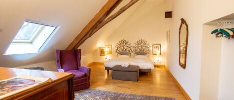 A cozy attic bedroom with a sloped ceiling, a wooden desk, a purple armchair, a bed with a headboard, and a small window.