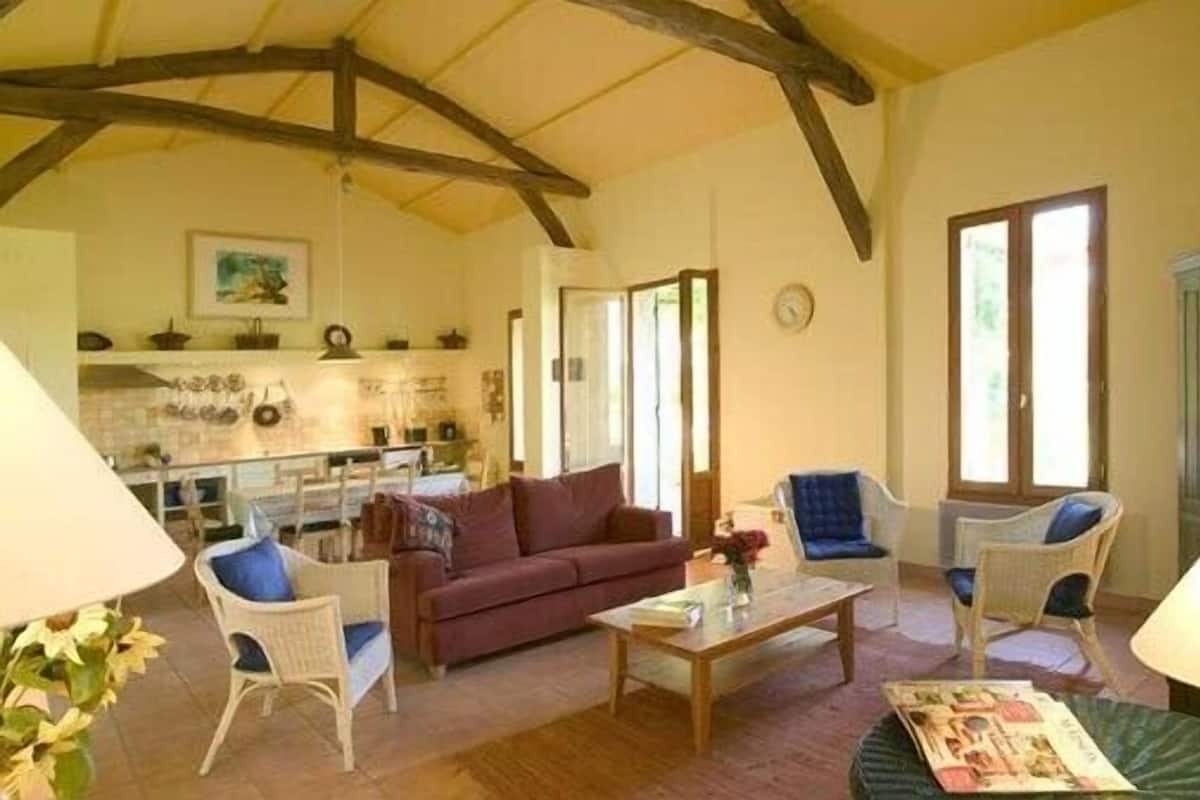 A cozy living room with a red sofa, white armchairs, and a wooden coffee table. A kitchen area with a window and a clock is visible in the background.