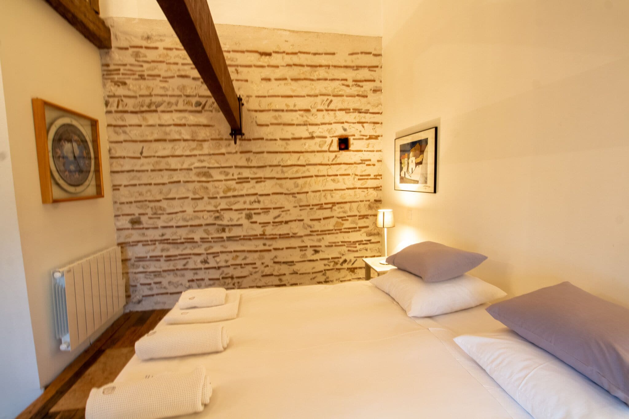 Un dormitorio con pared de ladrillo, una cama con sábanas blancas, una mesita de noche con lámpara y un cuadro enmarcado en la pared.