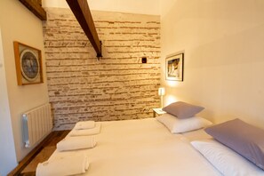 A bedroom with a brick wall, a bed with white linens, a nightstand with a lamp, and a framed picture on the wall. - Domaine Maison DoDo (Lamonzie-Saint-Martin)