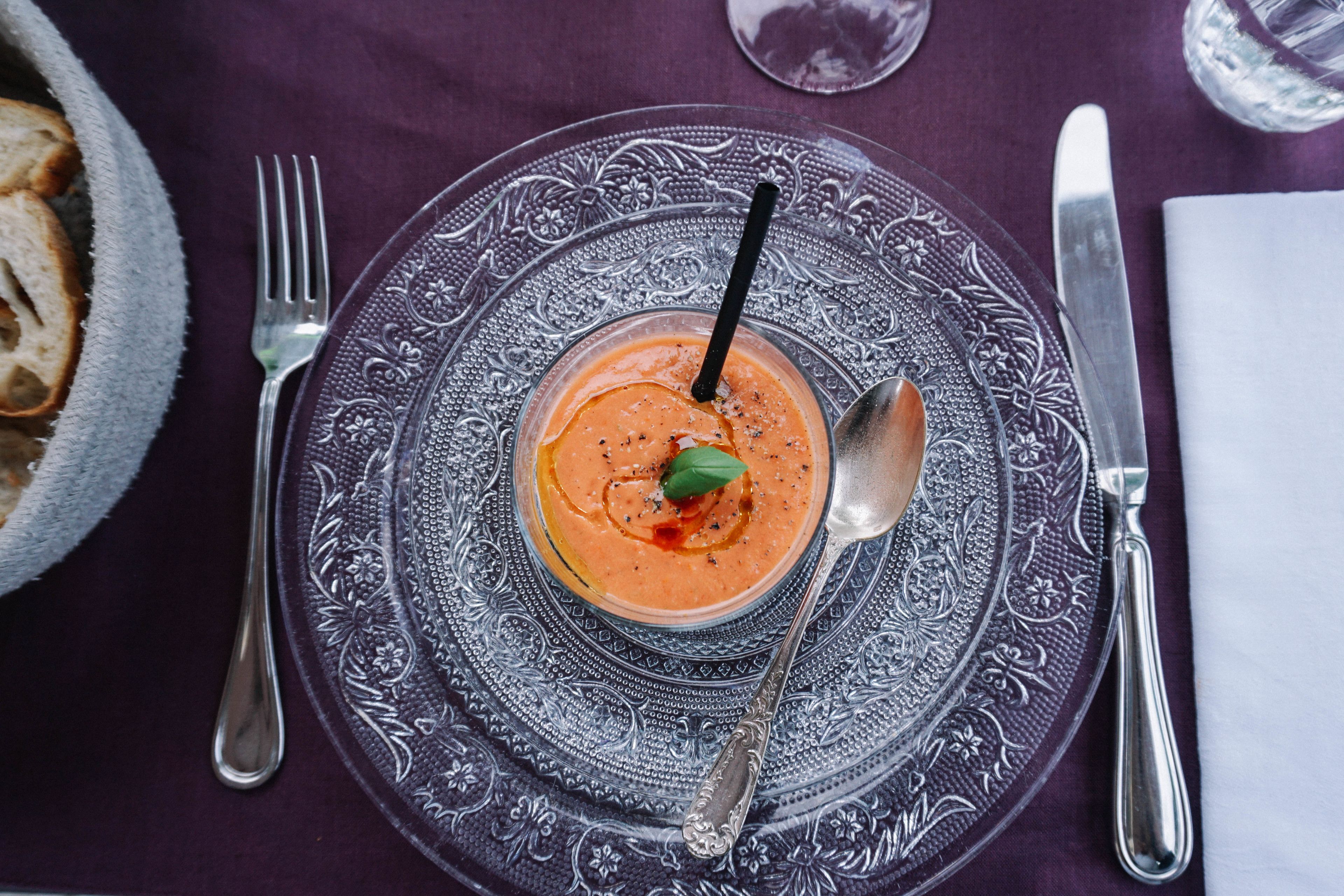 Un tazón de sopa sobre un plato ornamentado, con un tenedor y una cuchara a un lado, todo sobre un mantel.