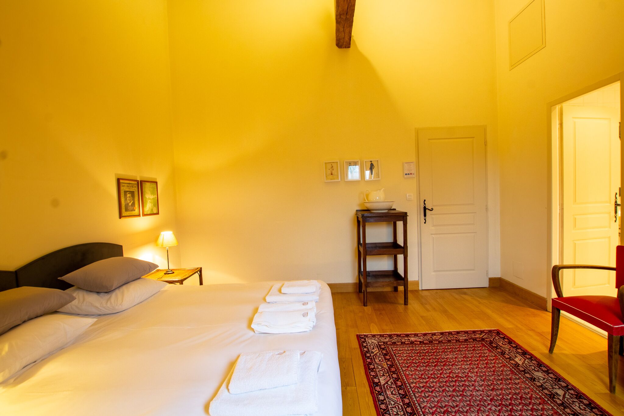 A hotel room with a bed, a nightstand, a red chair, a wooden door, and a small table with a bowl.