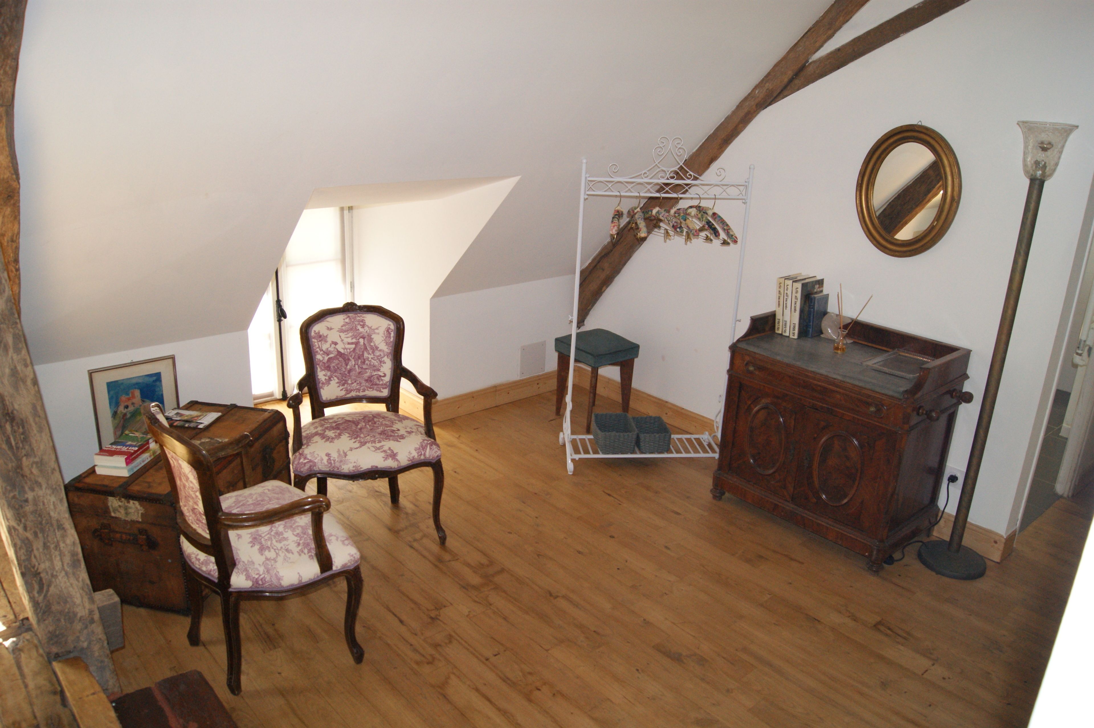 A room with wooden flooring, a wooden cabinet, two upholstered chairs, a small table with books, a mirror, and a tall floor lamp.