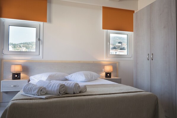 A hotel room with a bed, two bedside lamps, a nightstand, a wardrobe, and two windows with blinds. - Sirena Apartments (Malevizi)