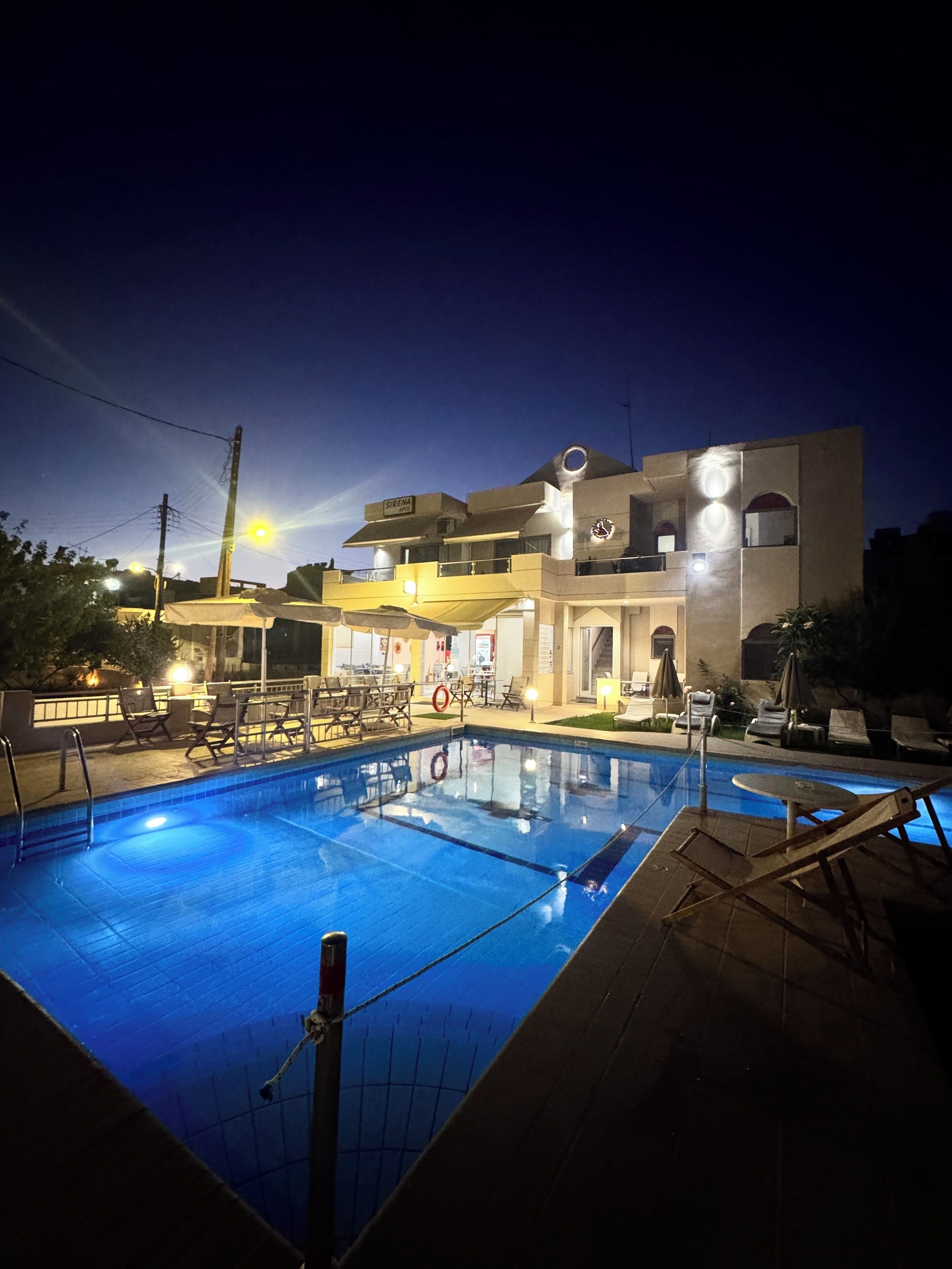 A swimming pool with lounge chairs and a building with a sign 'SIRENA APOTHEKE' in the background.