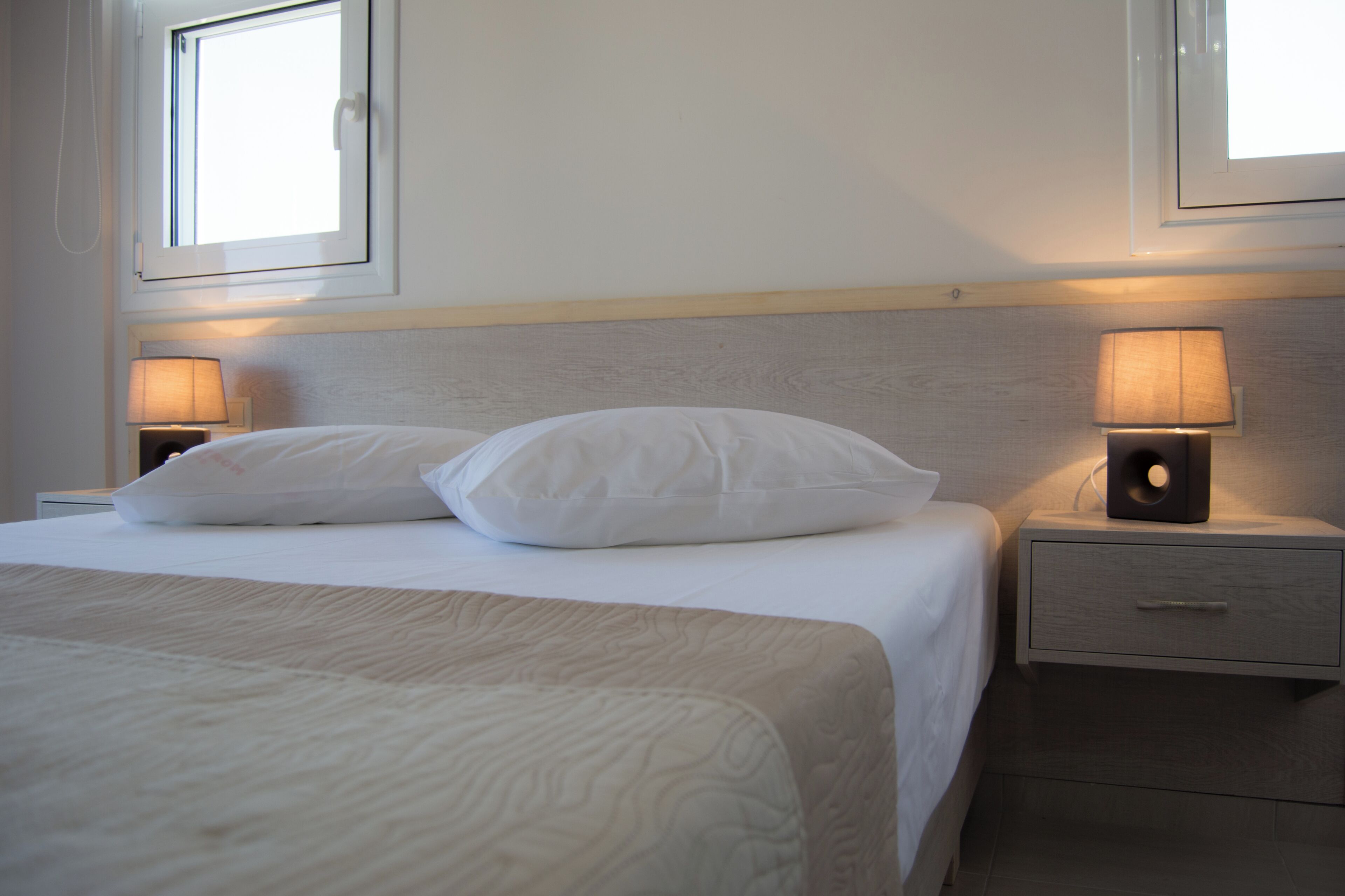 Ein ordentlich bezogenes Bett mit weißer Bettwäsche und Kissen, ein Nachttisch mit Lampe und eine Kommode mit Schublade.