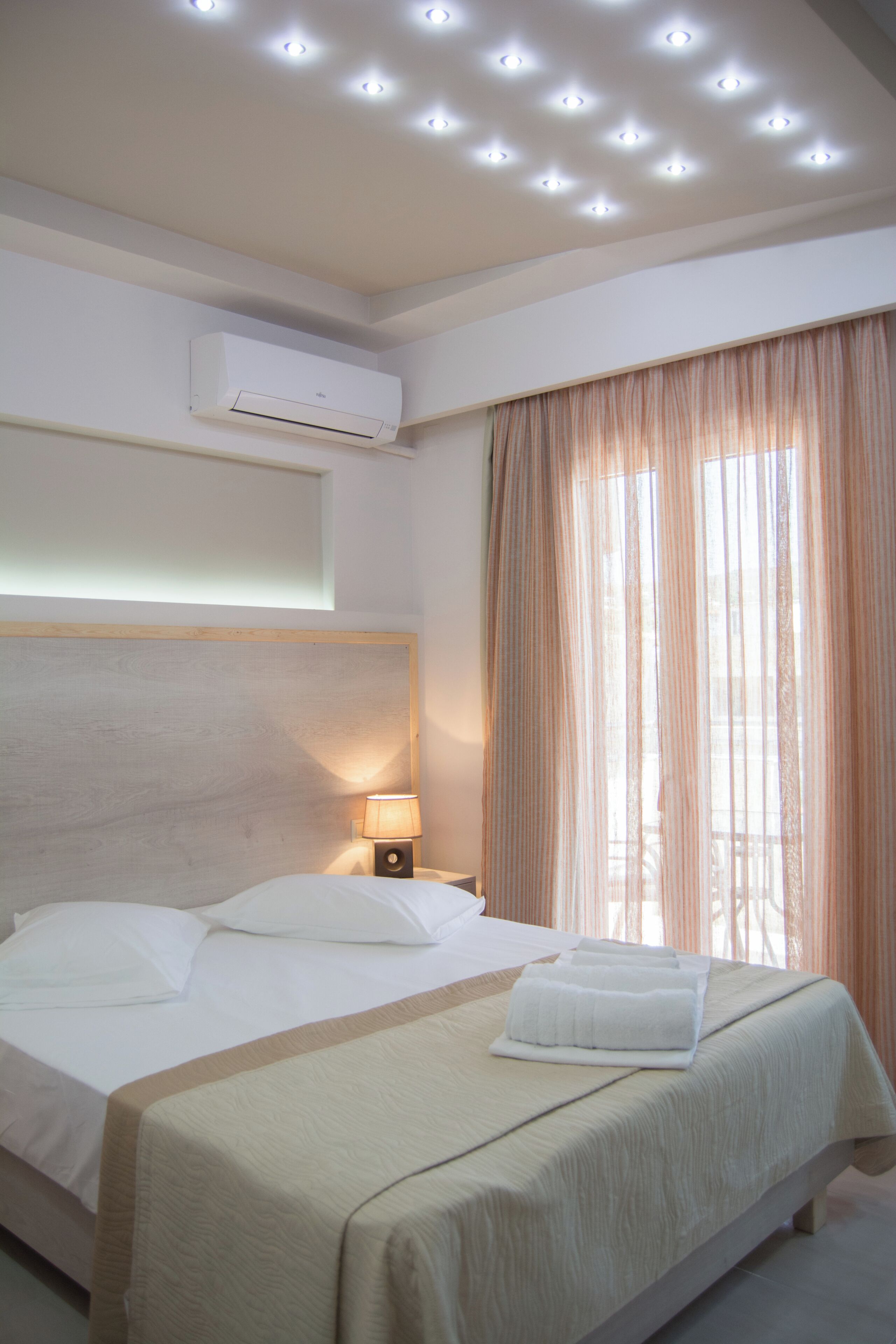 A hotel room with a bed, an air conditioning unit, a lamp, and a window with curtains.