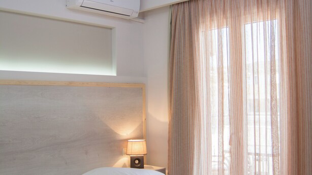 A hotel room with a bed, an air conditioning unit, a lamp, and a window with curtains. - Sirena Apartments (Malevizi)