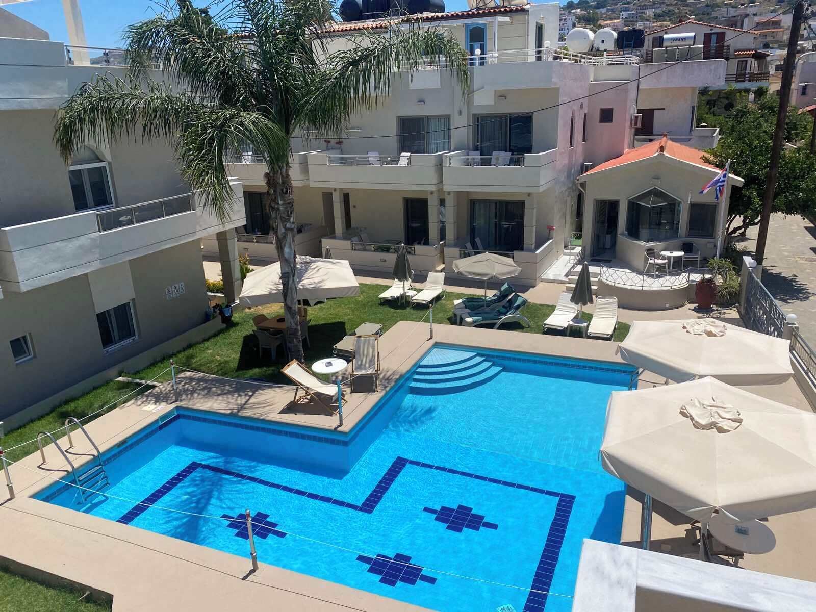 A swimming pool with a geometric tile pattern, surrounded by lounge chairs and umbrellas, with buildings in the background.