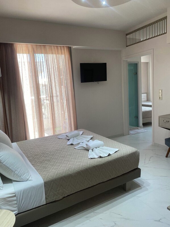 A hotel room with a bed, a flat-screen TV mounted on the wall, a bathroom visible through an open door, and a small desk with a chair.