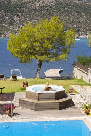 Un bain à remous où une personne est assise, entourée d’une piscine, d’un arbre et d’une vue sur un lac où se trouvent des bateaux.
