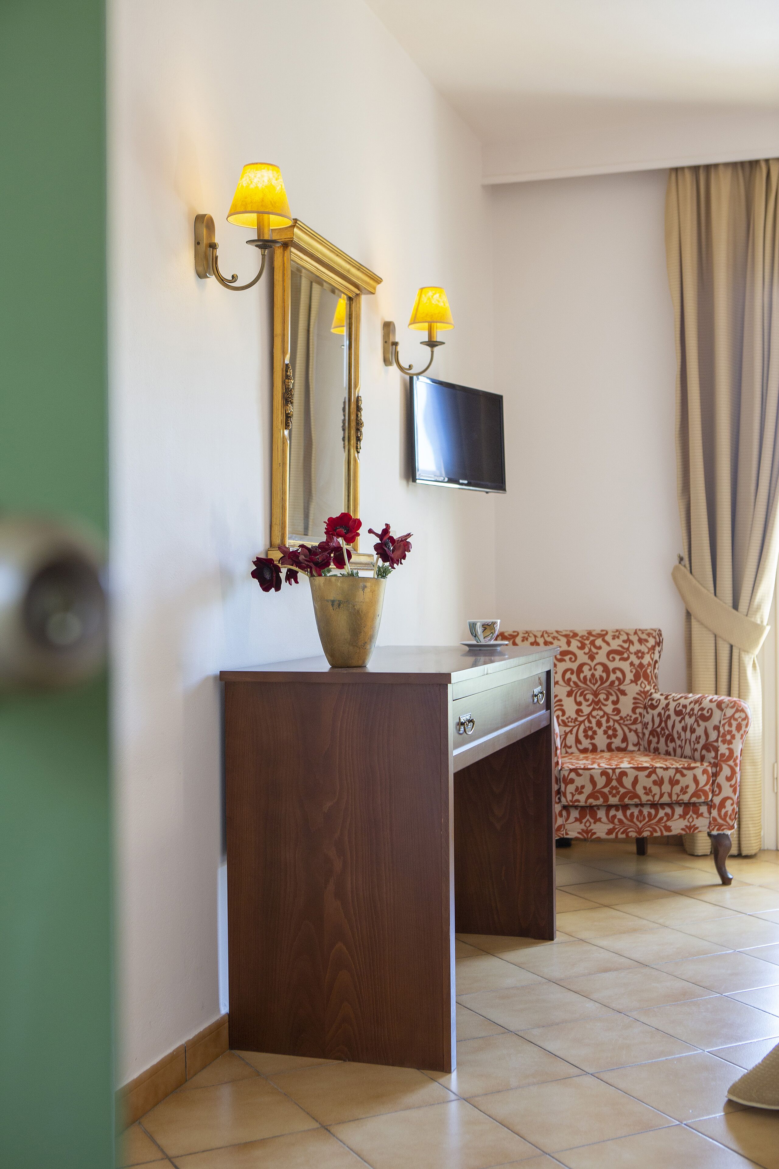 Une chambre d’hôtel avec une console en bois, un vase de fleurs, un miroir, un téléviseur à écran plat et un fauteuil à motifs.