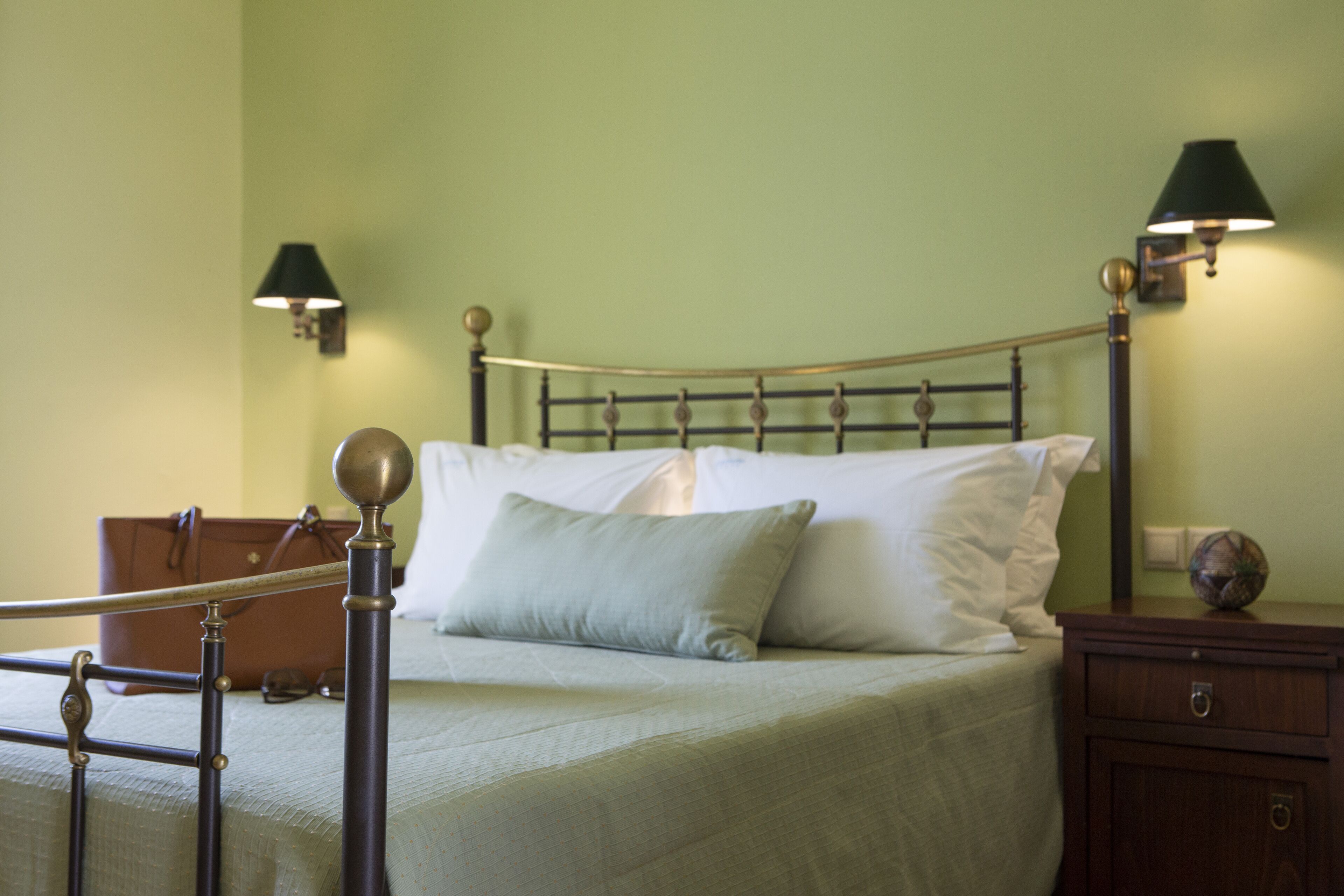 Een netjes opgemaakt bed met een metalen hoofdeinde, een nachtkastje met een lamp en een koffer op de vloer.
