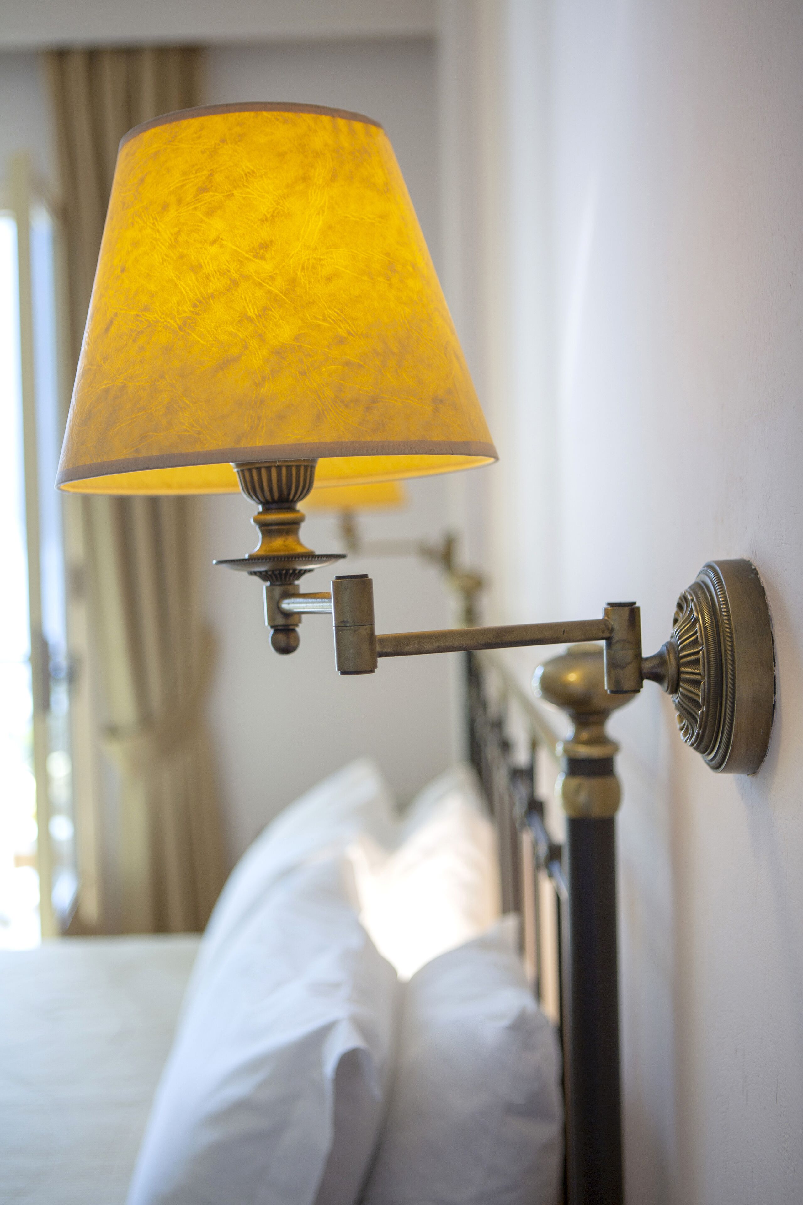 Una lampada a parete con un paralume giallo, un letto con lenzuola bianche e una finestra con tende.