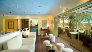 A spacious lounge area with a wooden floor, white sofas, and a glass wall offering a view of the outdoor patio. - Cleopatra Beach Hotel (Lefkada)
