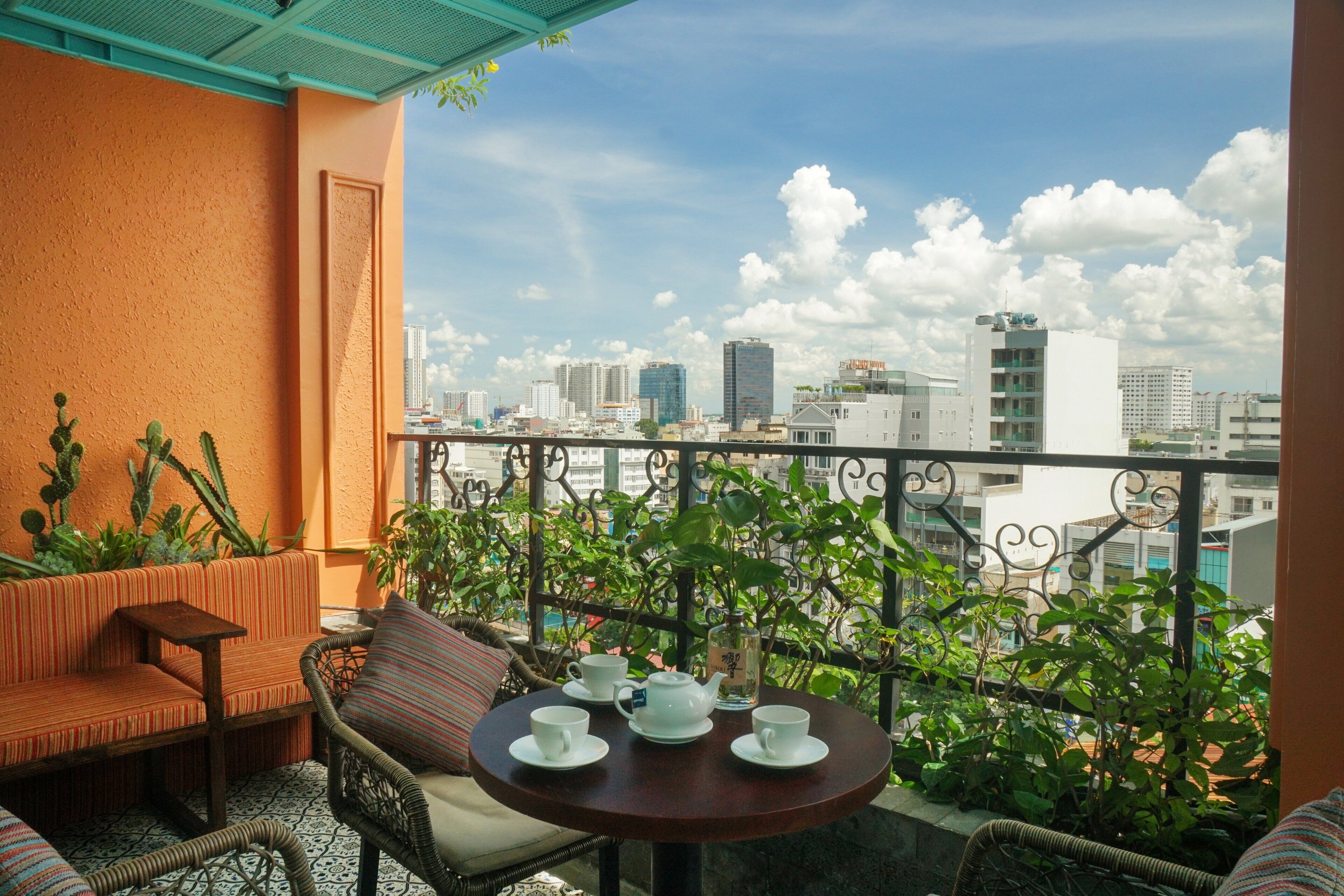 A balcony with a table set for tea, surrounded by wicker furniture and potted plants, overlooking a cityscape.