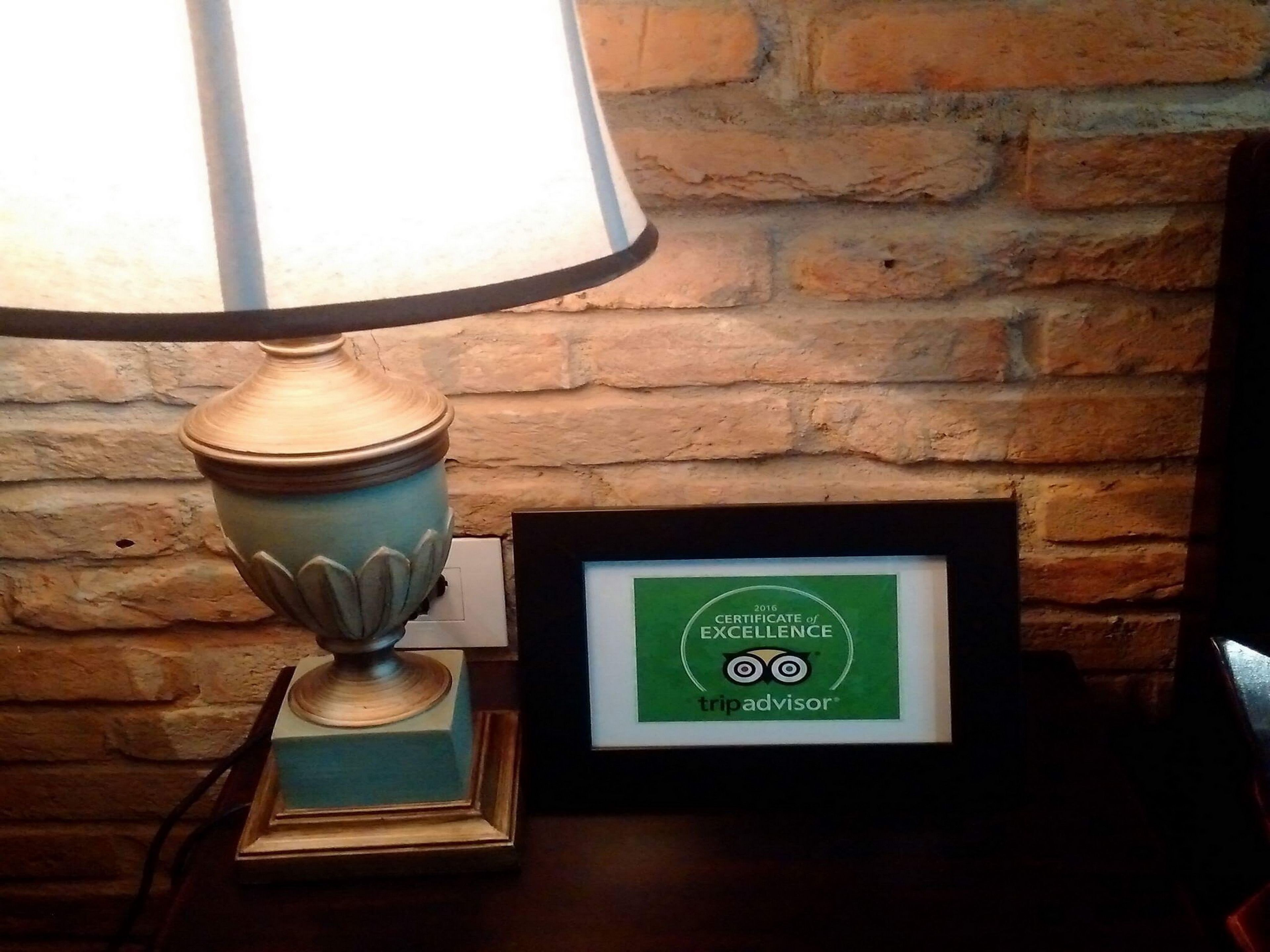 Uma luminária com cúpula azul sobre uma mesa, um certificado de excelência emoldurado e um fundo de parede de tijolos.
