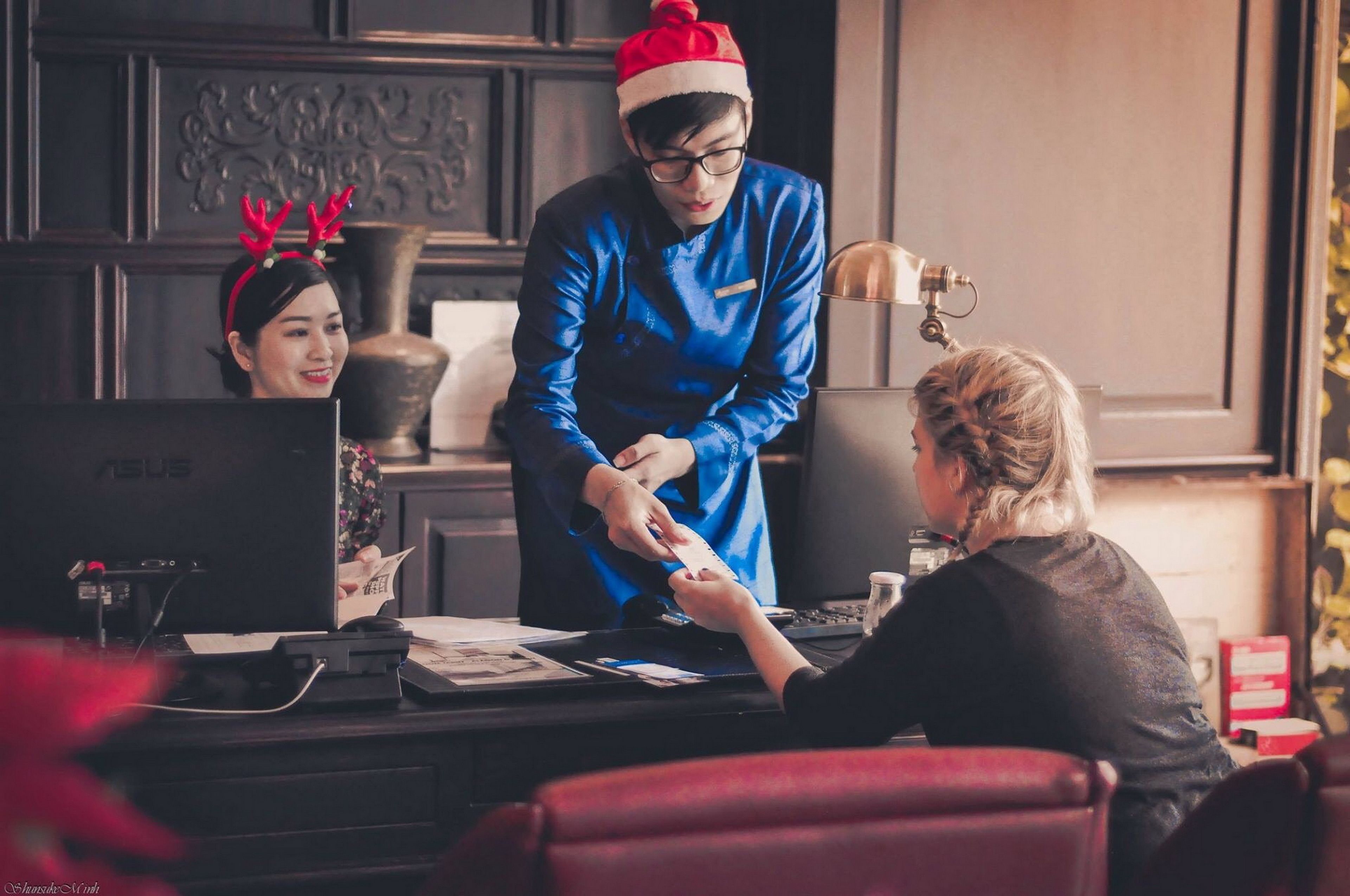 Ein Hotel-Empfangsbereich mit Theke, Computer und einer Person in blauer Uniform, die einer sitzenden Person eine Karte aushändigt.