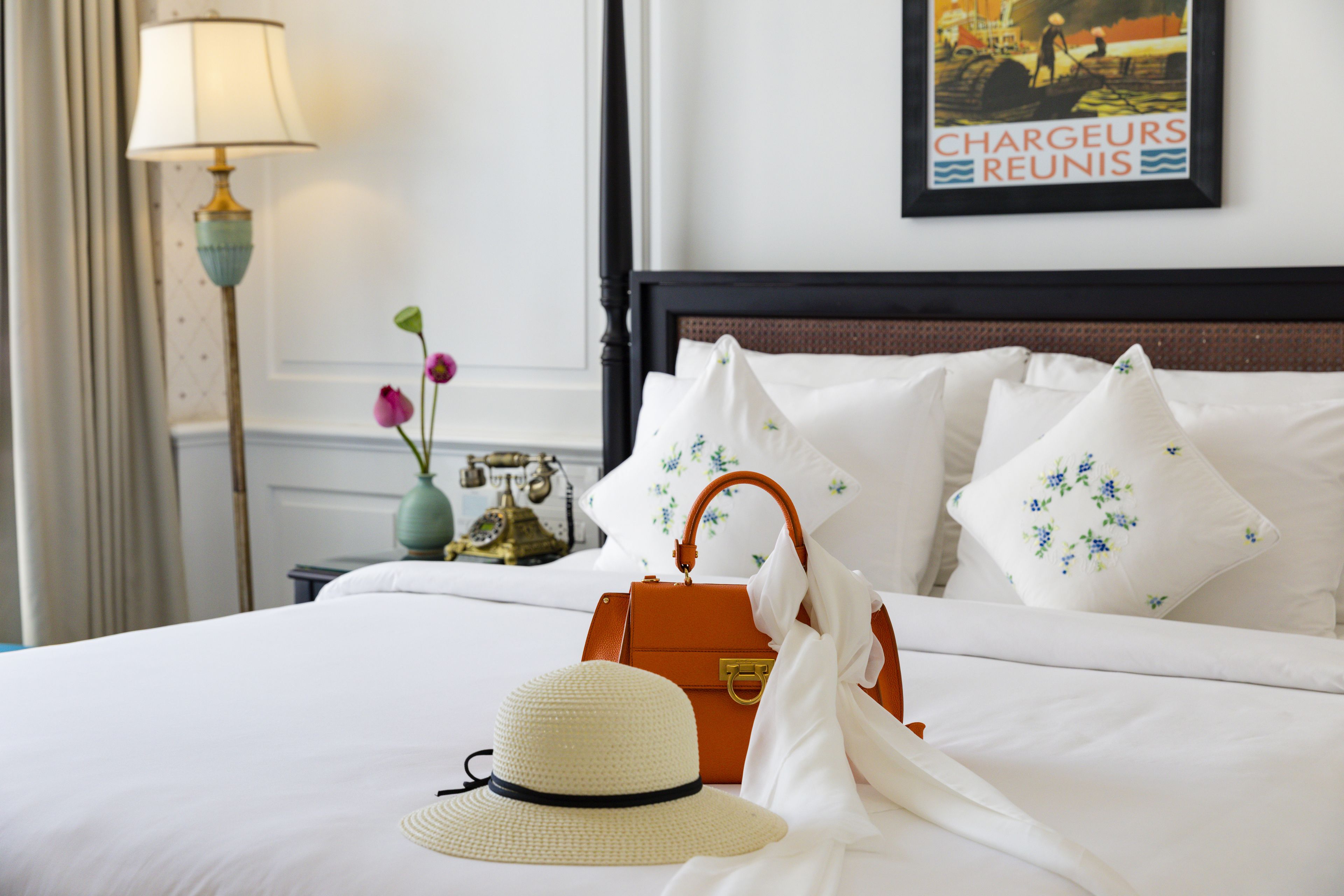 A neatly made bed with white linens, a floral pillow, a white hat, and an orange handbag.